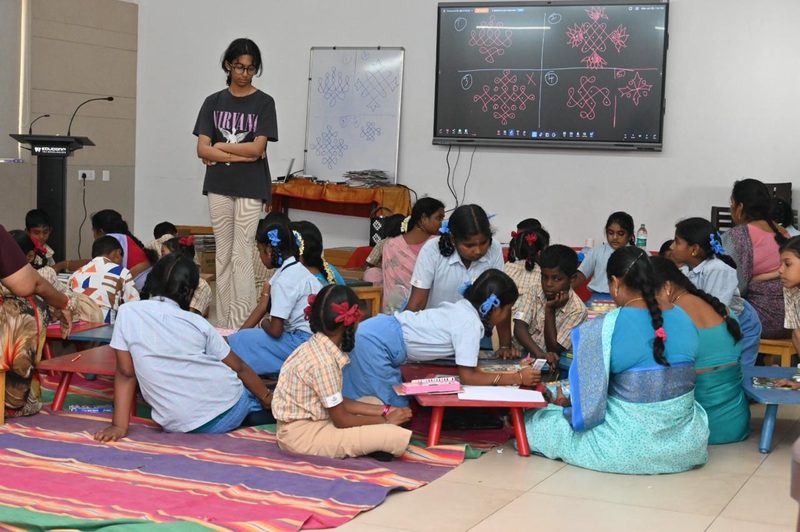 Children working on art activities on the floor while Shruti Arvind watches, with a kolam pattern on the screen behind